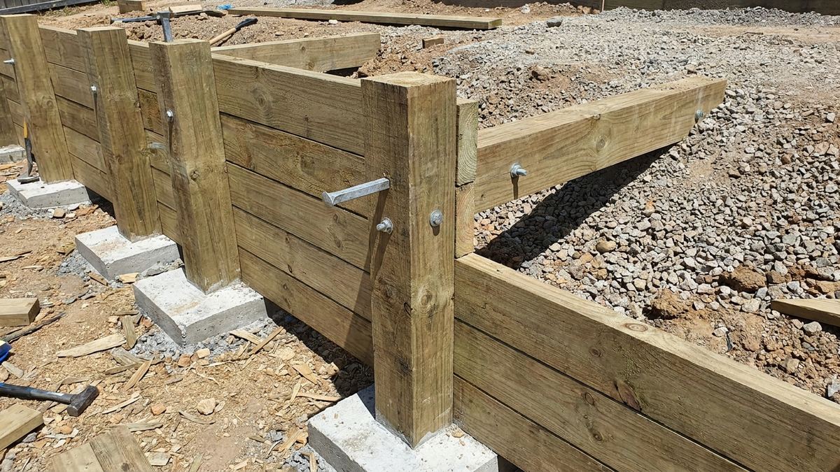 Pressure-treated timber retaining wall under construction with horizontal timbers between posts and a deadman tieback running back into gravel backfill